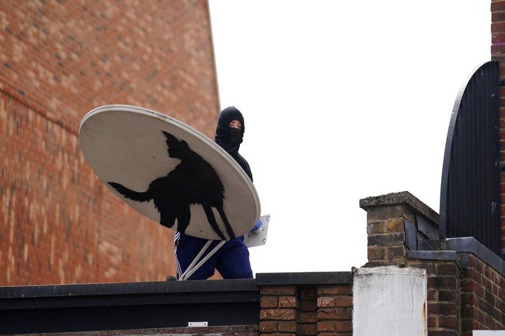 Una persona con el rostro cubierto retira una nueva obra de Banksy, que representa un lobo aullando pintado en una antena parabólica. (Jordan Pettitt/PA vía AP)